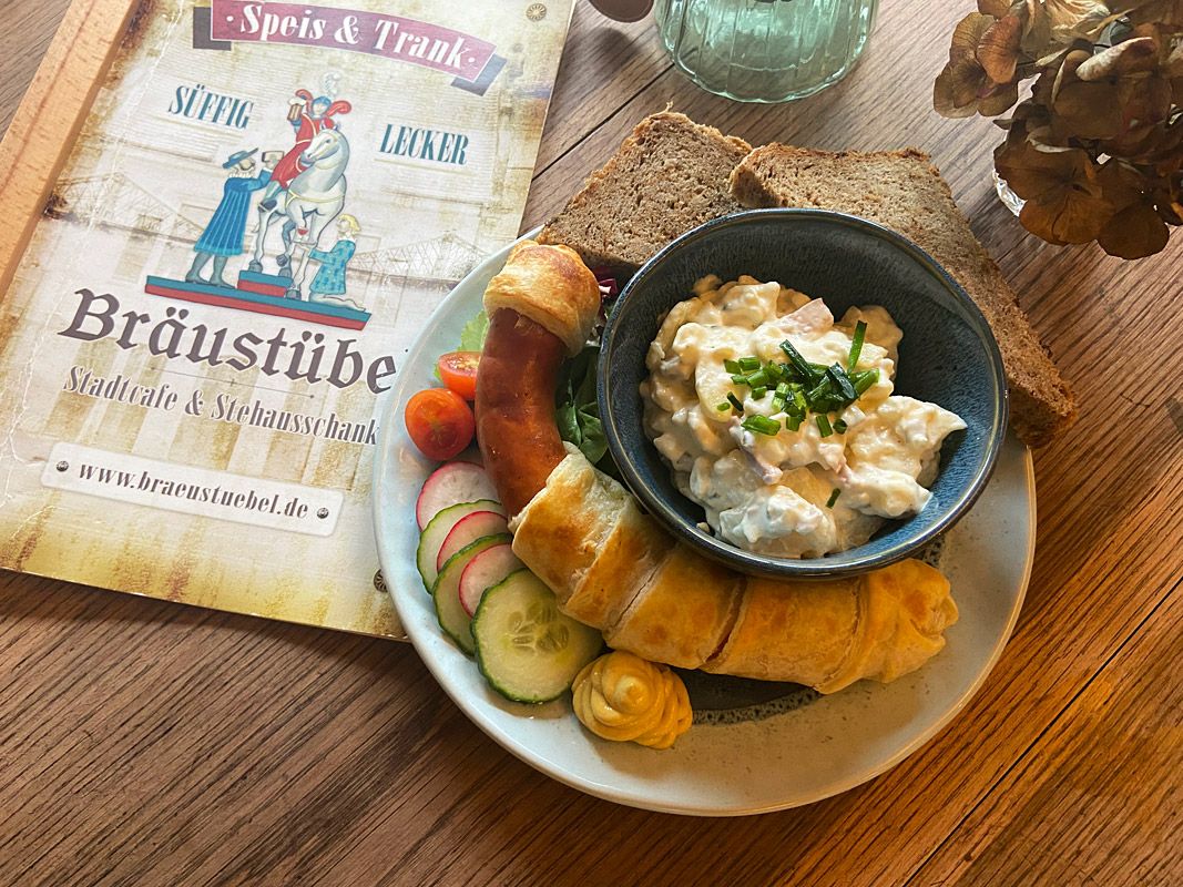 Wurst im Backmantel, Kartoffelsalat und Gemüse auf einem Teller präsentiert